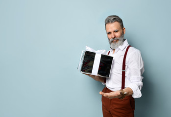 Wall Mural - Gray-haired man in white shirt, brown pants and suspenders. Smiling, holding silver gift box, posing against blue background