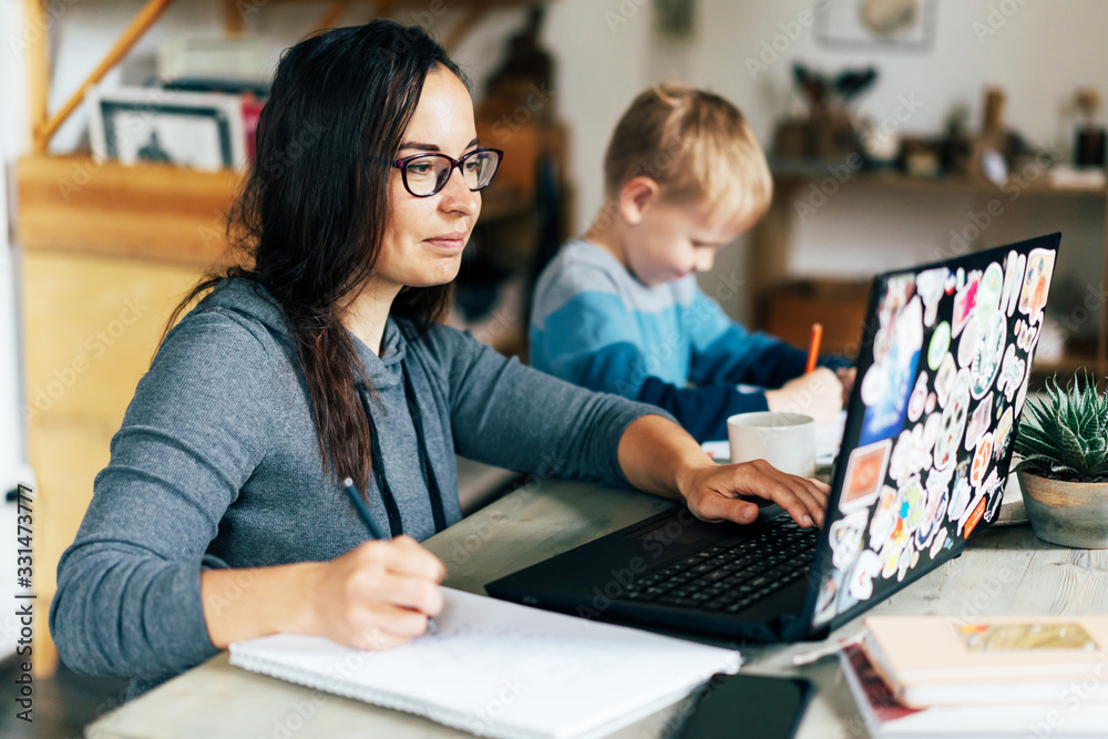 Concept of work from home and home family education. Mom and son are sitting at the desk. Business woman works on the Internet in a laptop, a child writes in a notebook. - obrazy, fototapety, plakaty 