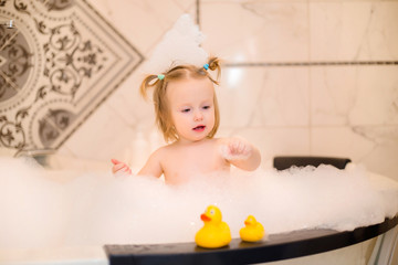 Wall Mural - Toddler girl bathes in bath with foam at home, plays with rubber duck