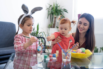 Happy young beautiful mother spending time with her joyful daughters. Cheerful family mom and children painting easter eggs with colors. Little pretty girl putting bunny ears. Preparation for Easter.