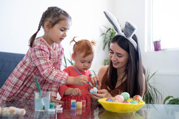 Happy young beautiful mother spending time with her joyful daughters. Cheerful family mom and children painting easter eggs with colors. Little pretty girl putting bunny ears. Preparation for Easter.