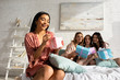 © LIGHTFIELD STUDIOS - Selective focus of african american girl opening present with friends holding gift boxes on bed at bachelorette party