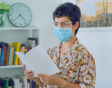A Woman Doing Research, A Young Woman Reads A Book While Wearing A Mask To Avoid Contagion, In The Background A Bookcase With A Clock