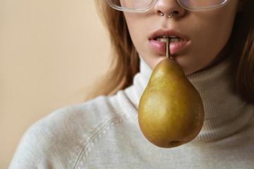 Wall Mural - Fashion sexy young lady hold pear in mouth, stylish girl with piercing in nose wear trendy glasses, vogue attractive model in eyewear with fruit on beige studio background, copy space, close up view