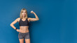 © Olga Rolencino - Portrait of beautiful athletic caucasian woman in sport bra and shorts showing biceps of her hand. Blue background. Copy space
