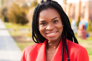 Wall Mural - Close up portrait of a beautiful young african american woman with pigtails in a red business suit smiling and walking along the street