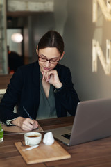 Business woman in a black suit and glasses sitting at a table in a cafe working at a laptop holding a notebook with records