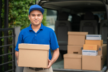 Wall Mural - A smiling young asian delivery in blue uniform with parcel cardboard in front of customer house. Messenger and delivery concept.