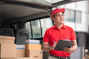 Wall Mural - A smiling young asian delivery in red uniform with parcel cardboard in front of customer house. Messenger and delivery concept.