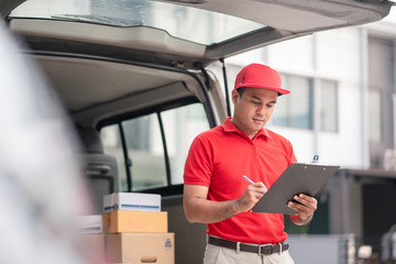 Wall Mural - A smiling young asian delivery in red uniform with parcel cardboard in front of customer house. Messenger and delivery concept.