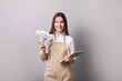 © LumenSt - Young woman in an apron with money on gray background. Barista concept