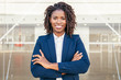 © Mangostar - Happy successful business leader posing near outside. Young business woman with arms folded standing near glass wall, looking at camera, smiling. African American businesswoman concept