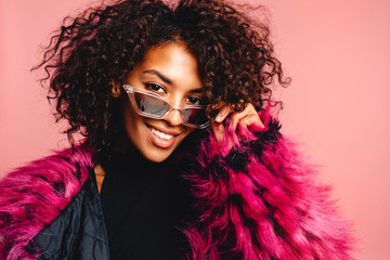 Studio photo of inspired african lady dancing with hands up. Indoor portrait of relaxed black girl isolated on pink background, enjoying party and wearing pink fur coat and white jeans. Horizontal.