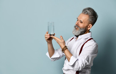 Wall Mural - Gray-haired man in years in white shirt and brown suspenders. He is showing glass of water, posing sideways on blue background