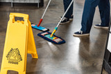 Fototapeta  - partial view of two cleaners washing floor with mops near wet floor caution sign