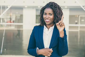 Wall Mural - Happy confident professional having great idea. Young African American business woman standing outside, pointing index finger up, looking at camera, smiling. Idea for startup concept