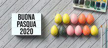 Easter Eggs With Message HAPPY EASTER 2020 In Italian And Lightbox With Number 2020 Surrounded By Brushes And Water Color Boxes On Wooden Background
