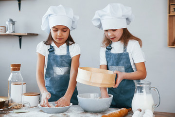 Two little girls in blue chef uniform working with flour by using sieve on the kitchen