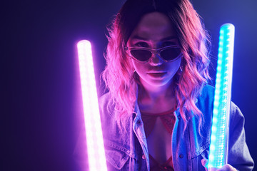 Wall Mural - Portrait of young girl in glasses holding light sticks in red and blue neon in studio