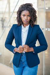 Wall Mural - Serious confident business lady posing near office building. Young black business woman standing at outdoor glass wall, adjusting jacket, looking at camera. Business portrait concept