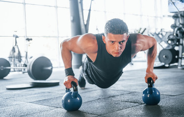 Wall Mural - Strong young man in sportive clothes doing push ups in gym by using dumbbells