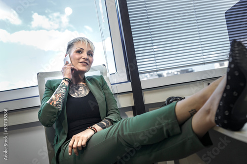 Fotografía Manager tatuata bionda, vestita con una giacca verde in ufficio , parla al cellulare seduta com eun boss nella sua poltrona