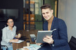 © AnnaStills - Young businessman holding tablet pc and using it for online communication with businesswoman sitting in the background at office