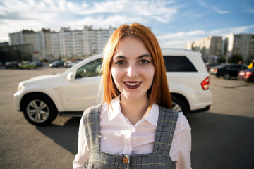 Wall Mural - Portrait of a fashionable teenage girl with red hair wearing casual dress standing outside in front of big new expensive car on warm summer day.