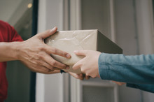 Hand Of Delivery Service Man In Red Uniform With Woman Customer Receiving Parcel Post Box From Courier At Home, Cargo Shipping, Fast Express Delivery Service, Online Shopping And Logistic Concept