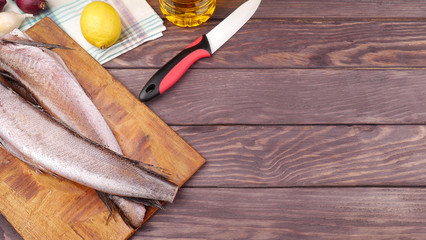 Two carcasses of fresh-frozen hake, pollock fish, on a cutting board on a wooden table. In the background is a knife, lemon, onion and a bottle of sunflower oil.