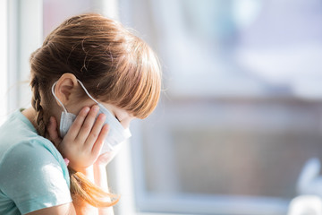 Adorable sad child girl by the window at home. Quarantine. Coronavirus. Epidemic.