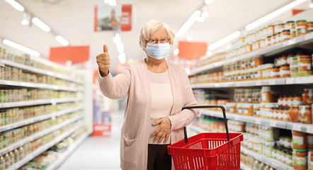 Wall Mural - Elderly woman shopping in a supermarket with a protective face mask showing thumbs up