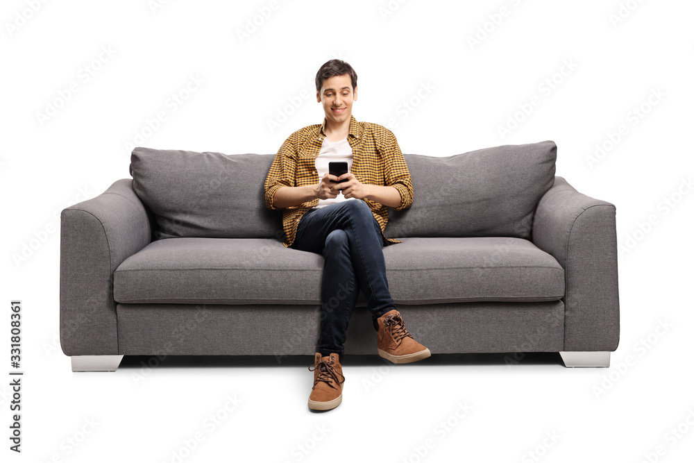 Happy young man sitting on a gray sofa and typing on a mobile phone - obrazy, fototapety, plakaty 