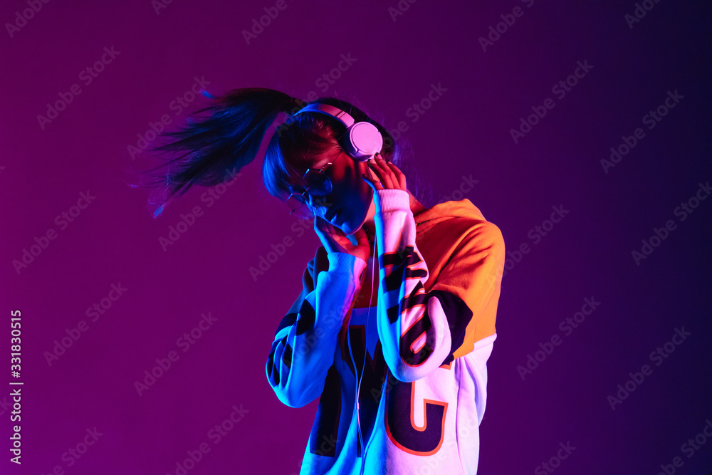 Stylish fashion teenager model wearing hoodie and headphones listening dj music dancing in purple neon lights. Young teen girl enjoy cool music 90s party mix in violet studio background. Copy space. - obrazy, fototapety, plakaty 