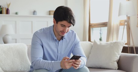 Wall Mural - Smiling millennial man using smart phone sitting on sofa in living room. Happy young male user texting sms message, playing game or chatting in internet social media dating mobile modern app at home.