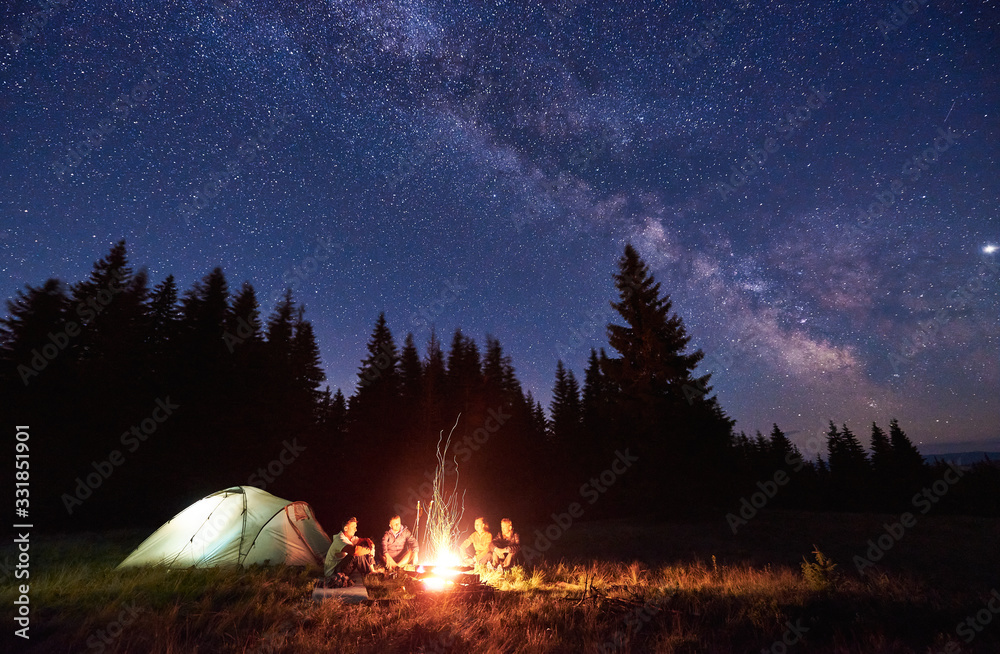 Night camping near bright fire in spruce forest under starry magical sky with milky way. Group of four friends sitting together around campfire, enjoying fresh air near tent. Tourism, camping concept. - obrazy, fototapety, plakaty 