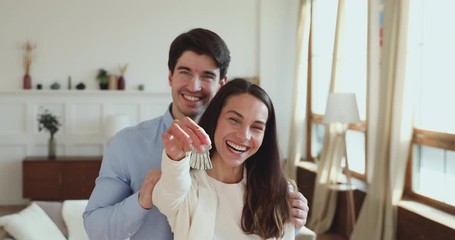 Wall Mural - Proud happy young adult couple holding keys standing in own new apartment. Smiling husband and wife first time home owners real estate buyers looking at camera. Family mortgage investment concept