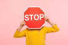 Unrecognizable Child In Yellow Sweatshirt Covering Face With Stop Symbol, Anonymous Person Holding Red Traffic Sign, Warning About Road Safety Rules. Indoor Studio Shot Isolated On Blue Background
