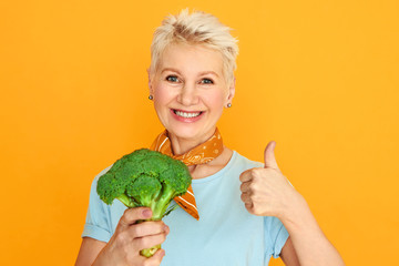 Attractive middle aged woman with short pixie hair holding fresh broccoli and smiling at camera choosing healthy organic food. Mature female making raw meal using vegetables, showing thumbs up