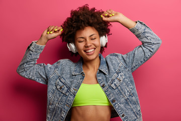Wall Mural - Positive hipster girl forgets about problems, raises arms, feels upbeat, dressed in stylish clothes, listens music in headphones, enjoys sound record, dances against pink wall, uses stereo accessory