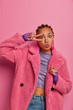 Portrait of fashionable ethnic woman makes peace victory sign near eye, keeps lips rounded, wears fashionable pink coat, purple sport gloves, poses stylish indoor. Body language. Fashion concept