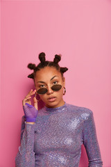 Self confident attractive female has funny hairstyle with buns, keeps hands on shades, wears glittering jumper, looks directly at camera, poses against pink pastel wall, empty space. People, style