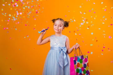 Wall Mural - happy little girl in a blue dress holds a gift bag and catches confetti while standing on a yellow background. holiday concept, space for text