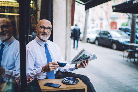 Aged male in glasses with cup of coffee and newspaper looking at camera