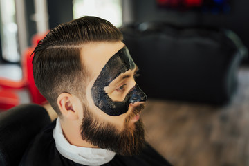 Handsome man with black charcoal face mask, concept skin care, pore cleansing from acne. Spa facial skin care for men, applying removal of black charcoal mask from nose face in barbershop.