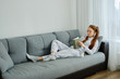 © bestsenny - A red-haired girl with glasses, with long tails in pajamas lies on the couch, turning over a book and looking down