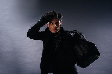 Canvas Print - Portrait of a young man with sharp jawline wearing a newsboy cap, holding a man bag while posing in studio on a white background.