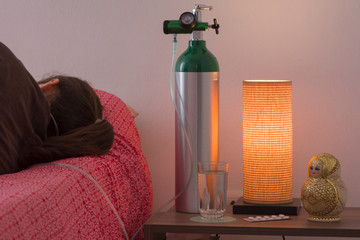 a woman backwards lies on her bed while uses a cannula connected to an oxygen cylinder at home