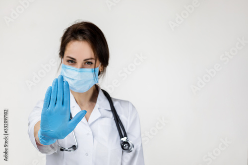 Fotografía Young woman doctor in mask and rubber gloves doing stop sign on gray background