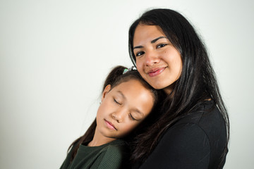 Little girl with her eyes closed rests on the arms of her mother who looks joyfully at the camera while hugging her little girl. Photography on white background